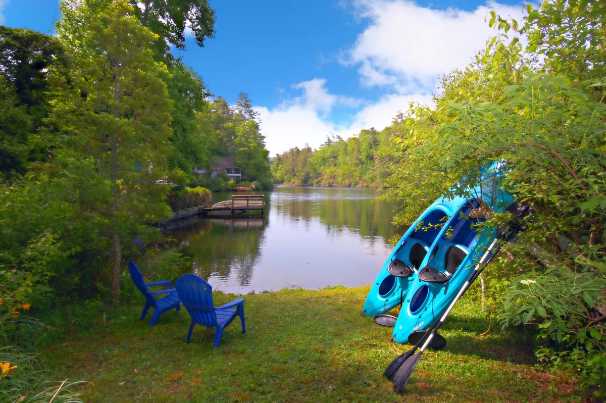 The image showcases a tranquil lakefront scene with calm water reflecting the sky, blue kayaks ready for adventure, inviting lawn chairs on a lush grassy area, and abundant greenery all around. The peaceful setting is ideal for outdoor activities like kayaking, fishing, or simply relaxing and soaking in the natural beauty. The image hints at memorable sunset views, opportunities to spot wildlife, and stargazing under clear night skies.
