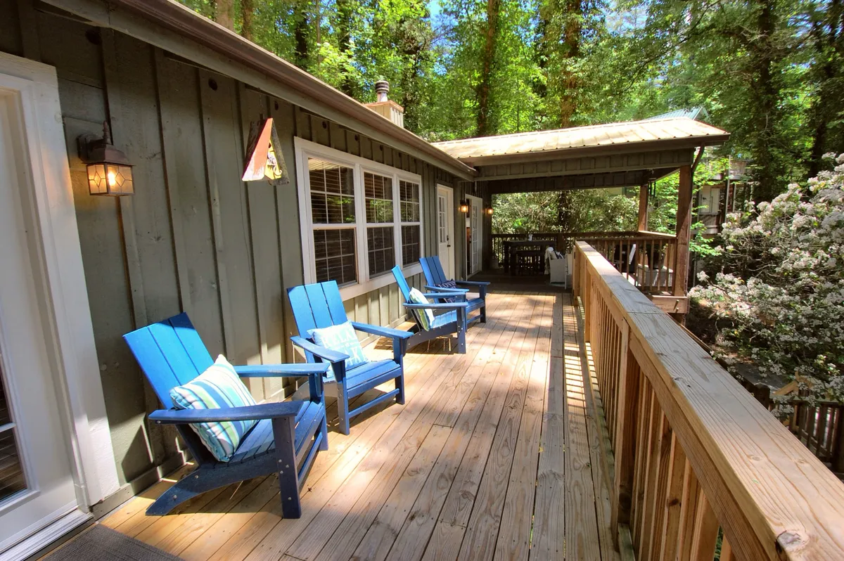 A peaceful wooden deck furnished with two blue Adirondack chairs, surrounded by vibrant green foliage. The deck is designed for relaxation, offering a serene spot to watch sunsets, listen to nature, or share quiet conversations, emphasizing comfort and connection with the outdoors.