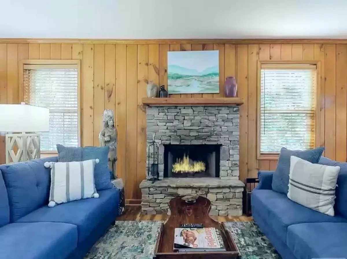 A warm living room with a stone fireplace, two blue sofas, wall art, and a central coffee table. The area feels perfect for gathering, relaxing, or working by the fire, with ample seating and a tranquil mood.