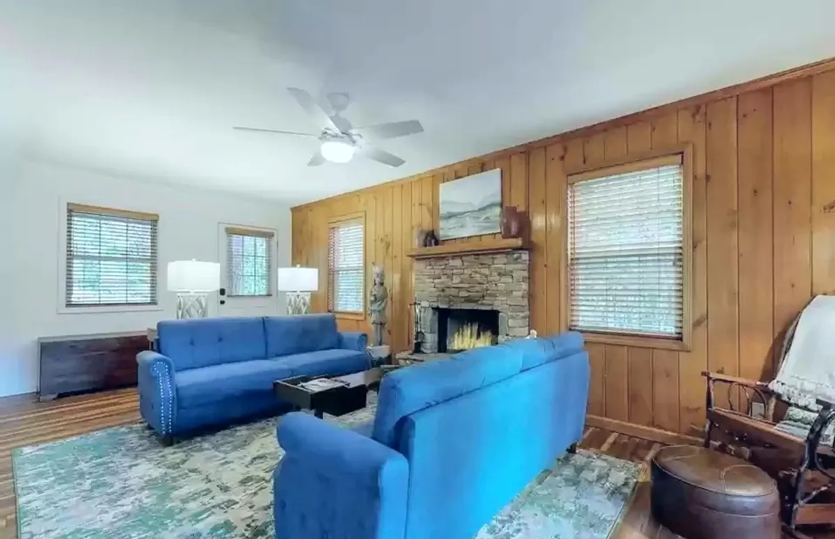A spacious living room featuring plush blue sofas, a striking stone fireplace as the focal point, rich wooden paneling, tasteful artwork above the mantle, and soft lighting from lamps on side tables. Sunlight streams in through large windows, enhancing the inviting, cozy ambiance.