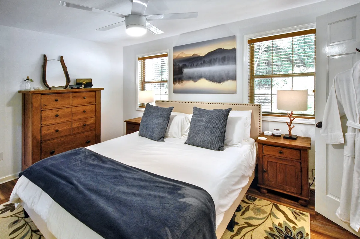 A restful bedroom scene with a comfortable bed adorned with a gray throw blanket, a wooden dresser, tasteful artwork above the bed, and lamps on each nightstand. The inviting setup speaks to a peaceful, cozy atmosphere ideal for relaxation or a good night's sleep.