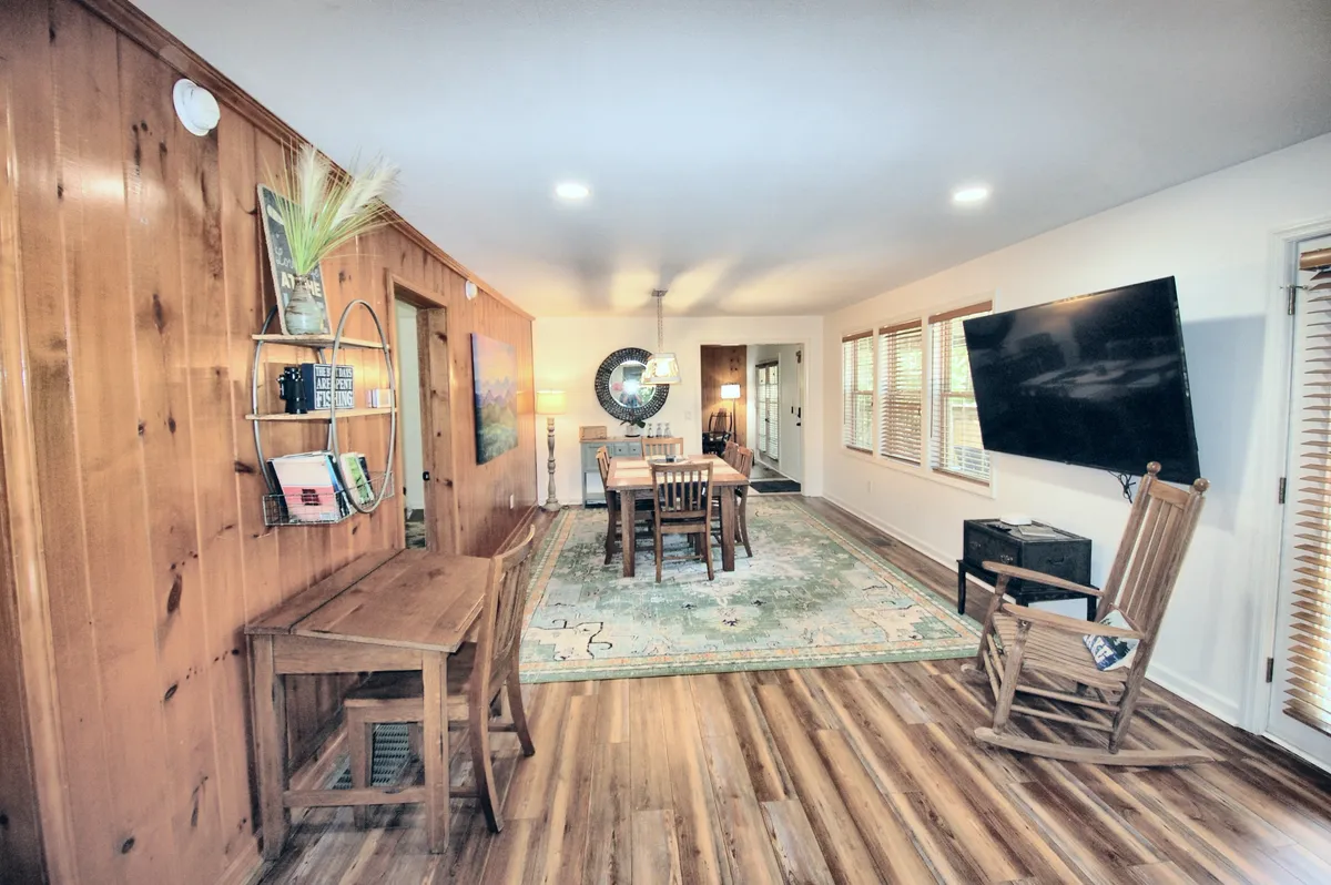 A warm and inviting dining space with a wooden table at the center, surrounded by comfortable chairs and anchored by a decorative rug. The room features wooden wall paneling, a rocking chair, and a prominent large smart TV, ideal for group gatherings and shared experiences.