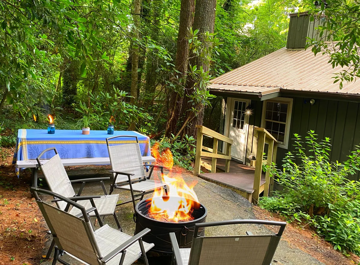 This exterior image showcases a fire pit surrounded by comfortable outdoor seating and a table set for dining, nestled beneath tall trees. The ambiance is intimate and warm, perfect for couples to enjoy an evening by the fire, toast marshmallows, and dine under the stars.