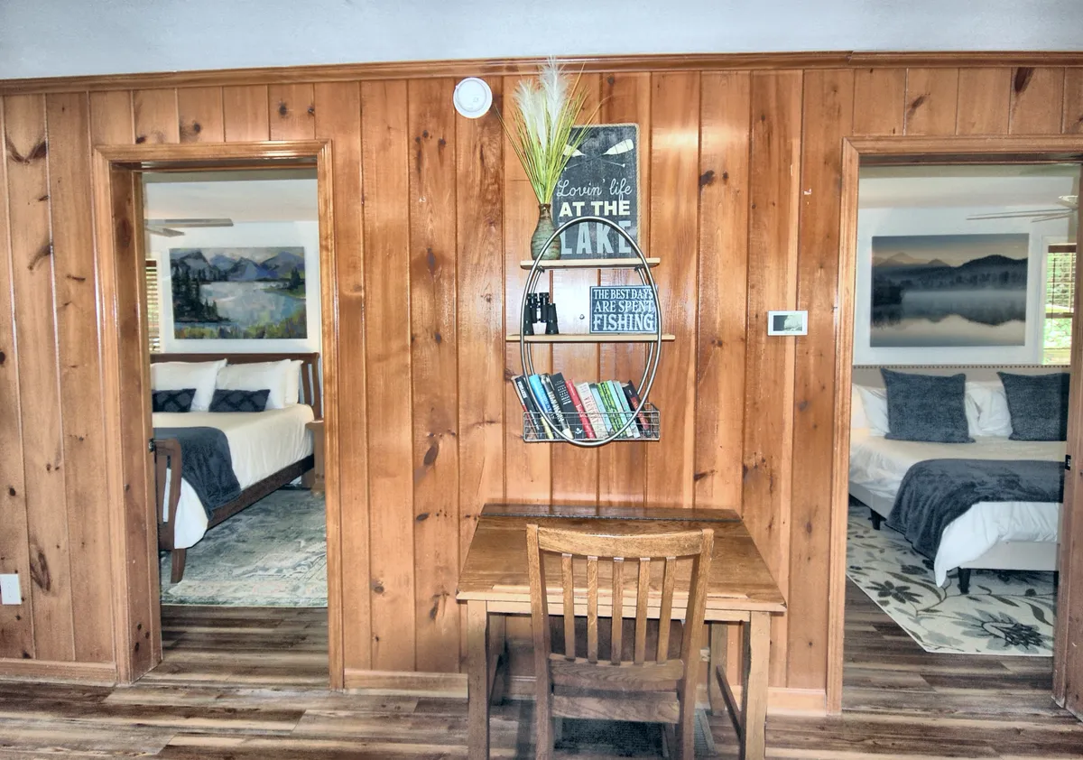 An interior corner with wood paneling, a decorative shelf stocked with books and a small plant, and two doorways leading to bedrooms. The setting is calm and encourages quiet reading or family game time.