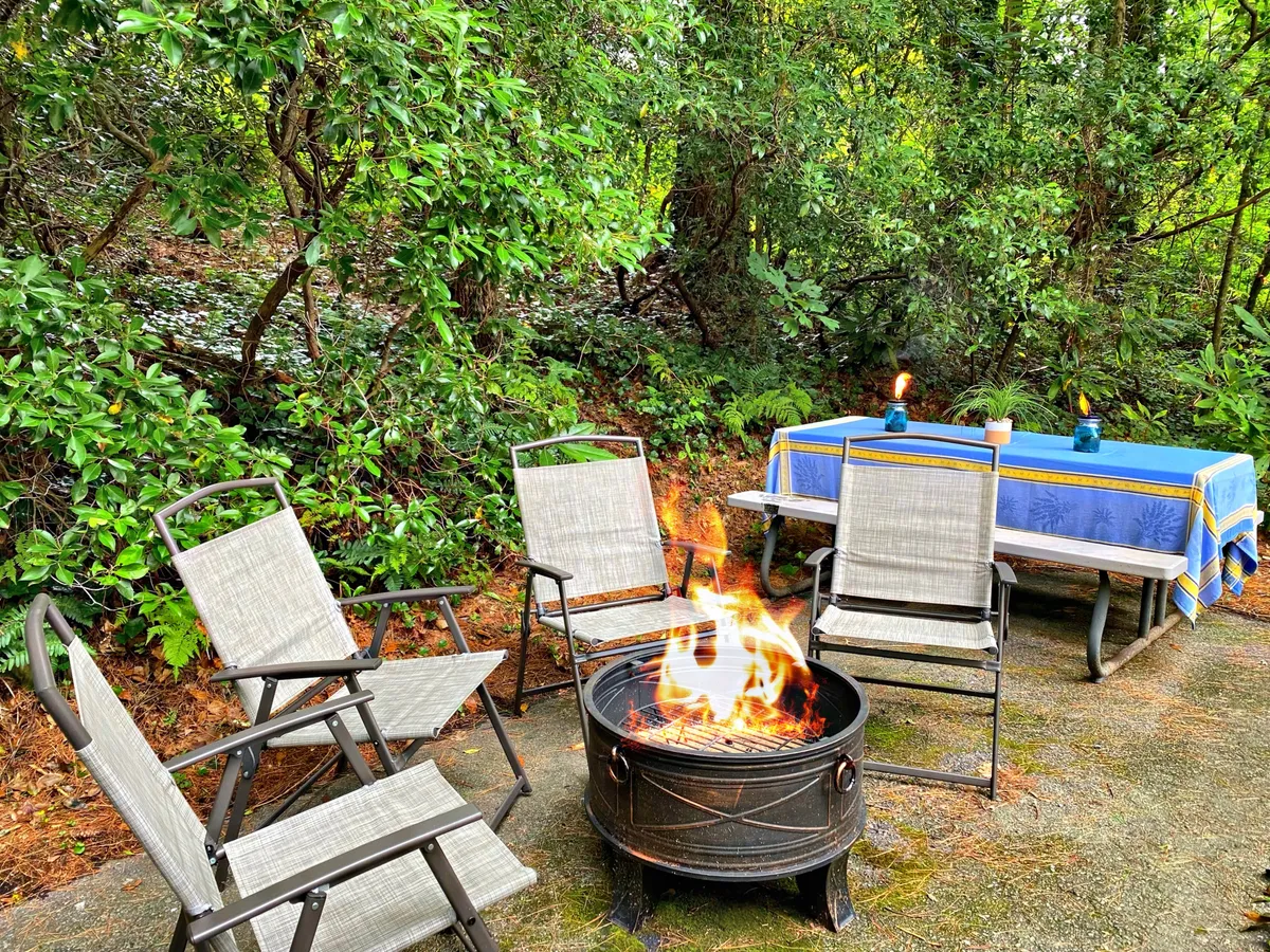 An inviting outdoor space featuring a fire pit, comfortable seating, and a table covered with a blue tablecloth. The area is surrounded by greenery, providing a relaxing backdrop for gatherings, stargazing, or unwinding after a productive workday.