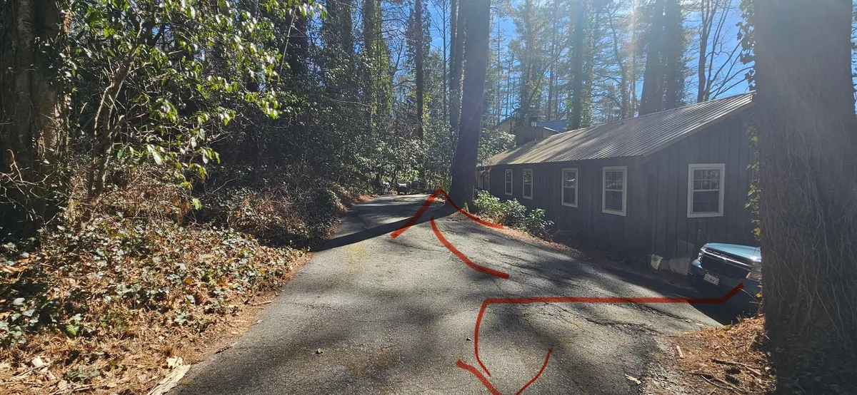 A scenic exterior shot featuring the home's driveway framed by lush trees and highlighting the inviting, cabin-like structure at the edge of the property. The wooden porch and natural surroundings evoke the peaceful seclusion of a mountain retreat, making it ideal for relaxing or connecting with nature year-round.
