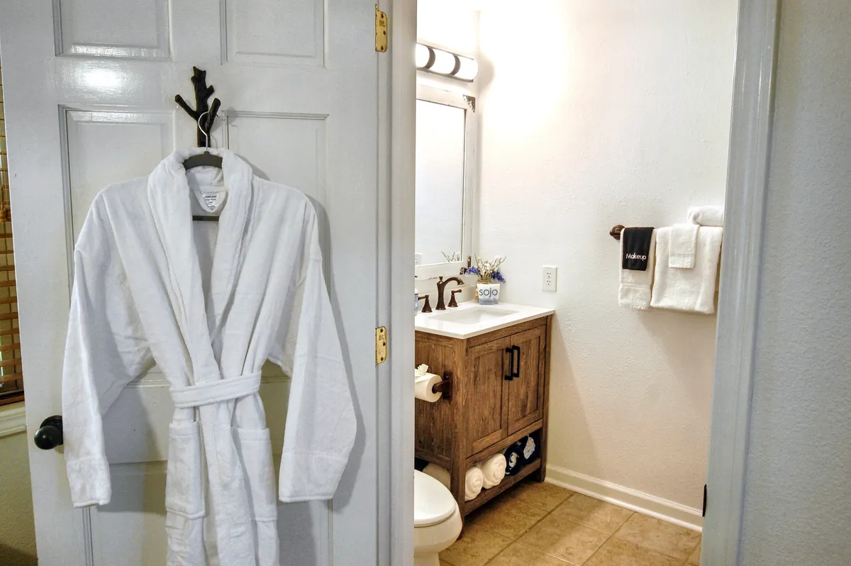 A spa-inspired bathroom with a pristine white bathrobe hanging by a wooden vanity, a mirror, and neatly folded towels. The space feels inviting and luxurious, ideal for unwinding and self-care routines.