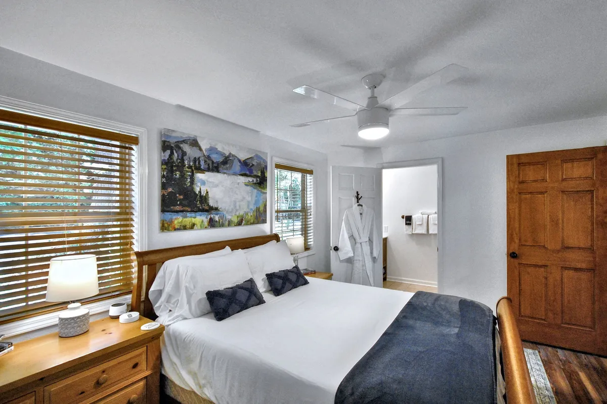 A tranquil bedroom featuring a neatly made bed, soft artwork above the headboard, large windows inviting natural light, and a door leading to an en-suite bathroom. The neutral color palette and uncluttered space evoke a sense of calm and relaxation.